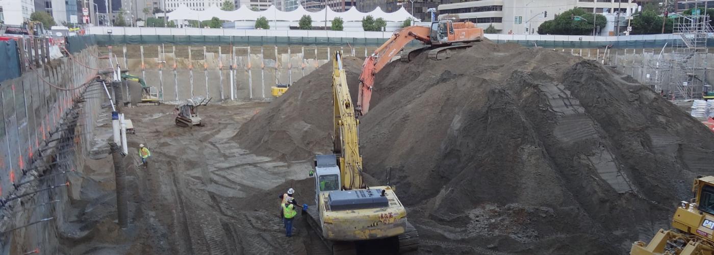 Helmeted construction workers and two large excavators inside active construction site pit.