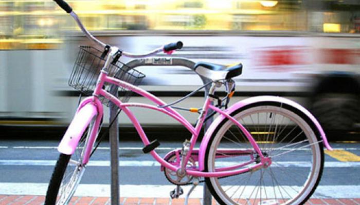 A close-up view of a pink bicycle with a front wire basket parked at a metal rack on a city sidewalk while a blurred vehicle passes by in the background.