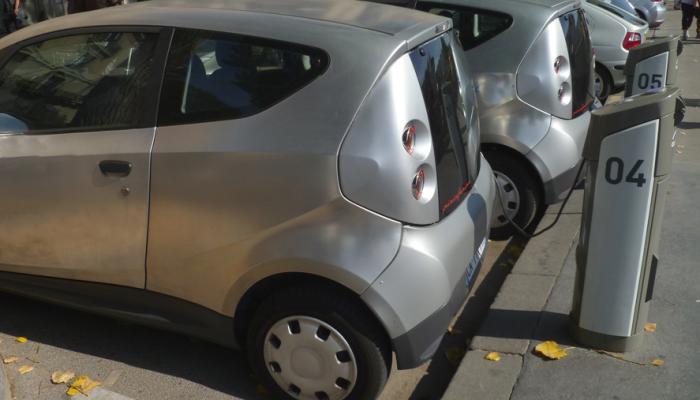 A row of several small, silver electric cars parked at a curbside charging station with designated gray charging kiosks numbered four and five in an urban setting.