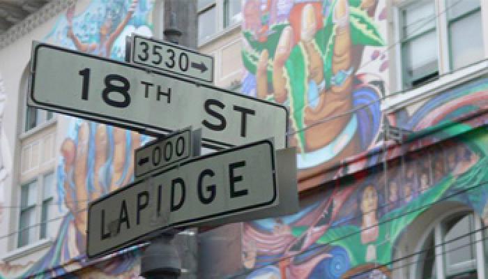 Mural on a building facade at the intersection of 18th Street and Lapidge Street, featuring colorful artwork and a directional street sign.