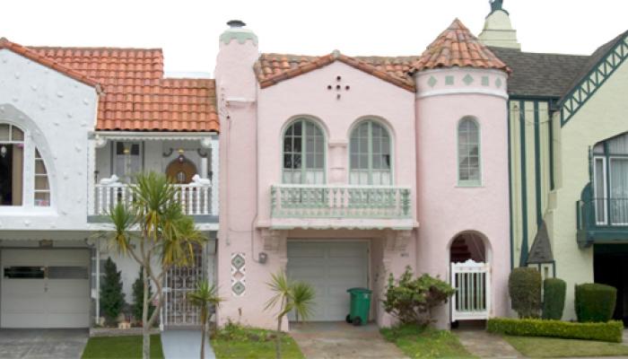 A row of three colorful, attached Mediterranean-style houses featuring pastel pink and beige facades, ornate arched windows, and decorative rooflines under a clear sky.
