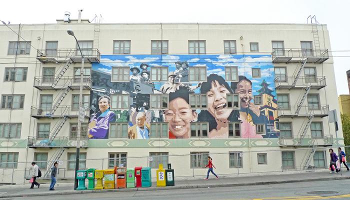 Building in Chinatown featuring a large mural of smiling children and community members, with people walking on the sidewalk below.