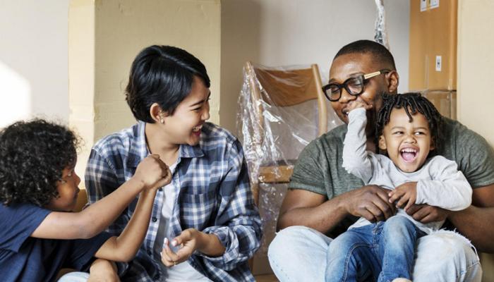 A happy family of three—a mother, father, and young child—celebrating in their new home. They are surrounded by cardboard moving boxes, with the father holding the child and everyone laughing together.