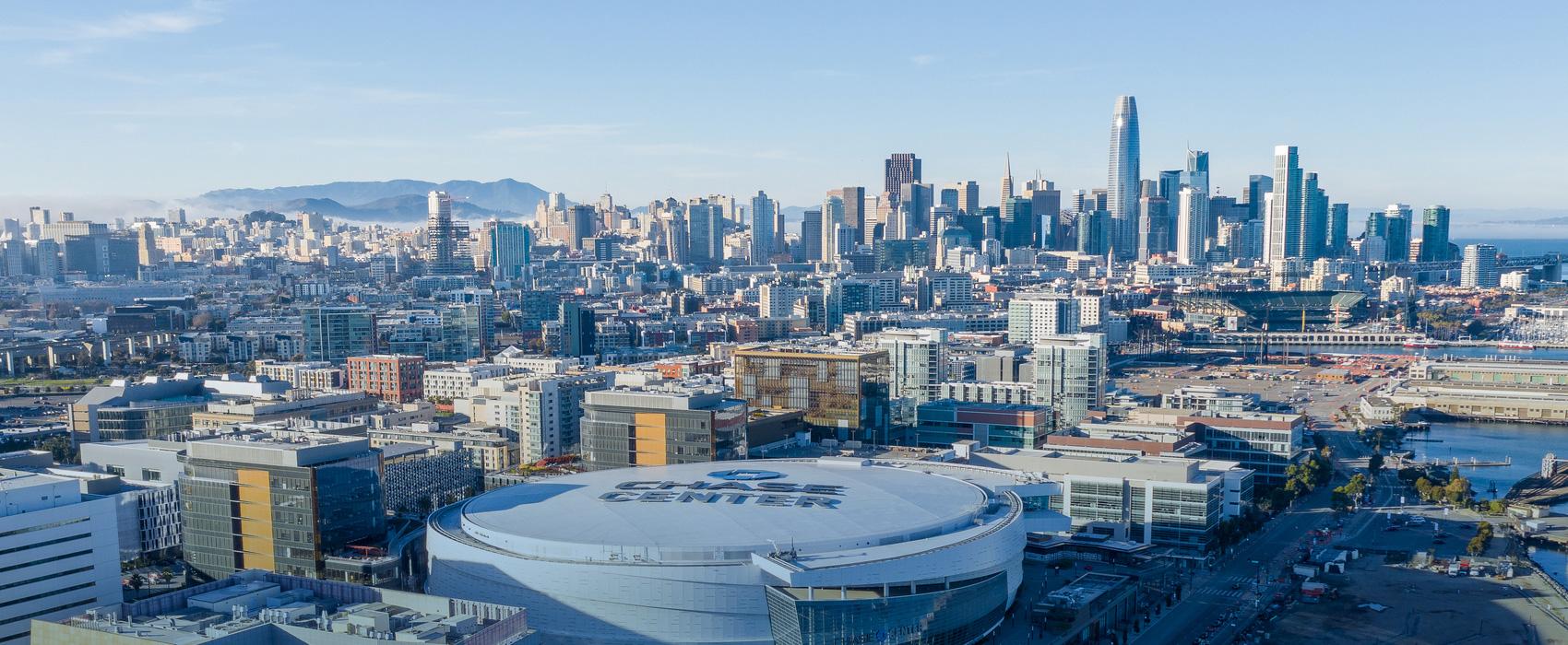 A panoramic, wide-angle view of the San Francisco city skyline at dusk with modern high-rise buildings and skyscrapers silhouetted against a hazy, light-blue sky.