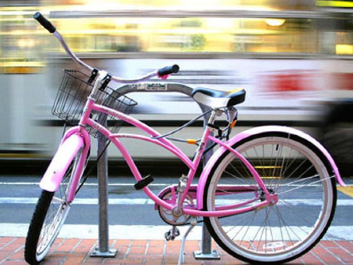 A close-up view of a pink bicycle with a front wire basket parked at a metal rack on a city sidewalk while a blurred vehicle passes by in the background.