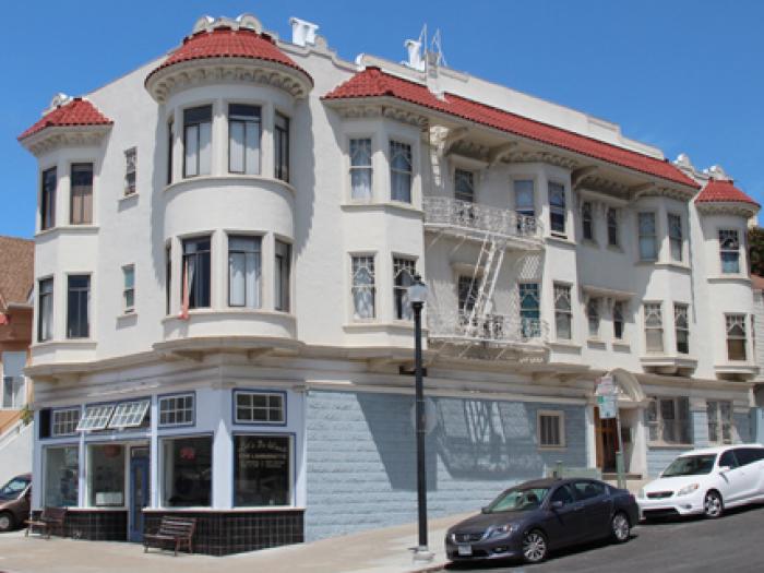 A corner-view of a multi-story, classic Victorian-style building in San Francisco featuring a curved facade with white bay windows, light beige siding, and intricate architectural detailing under a clear blue sky.