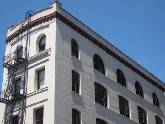 A low-angle, corner view of a tall, grey industrial-style building featuring a grid of rectangular windows and a black metal fire escape extending down the side.