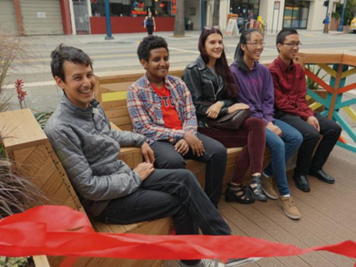 A group of five smiling young adults sits together on a wooden outdoor bench. They are dressed in casual sweaters and jackets, posed closely for a portrait in a vibrant public seating area.