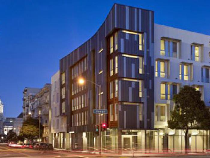 An architectural photograph of the modern residential building at 365 Fulton Street at night, featuring illuminated windows and a glowing ground-level storefront, with the lit dome of San Francisco City Hall visible in the background.