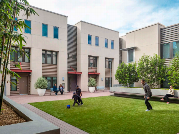 A modern, multi-story residential building with light gray and wood-toned siding overlooks a green courtyard where a person is walking and another is seated on a bench.