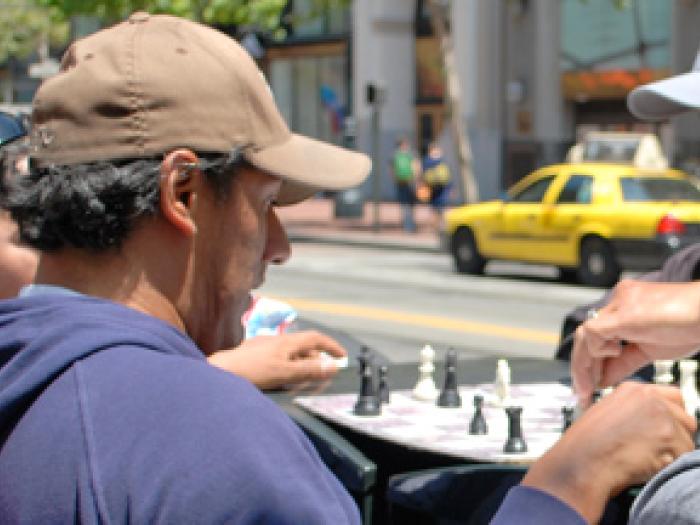 Two men and a child sitting at an outdoor table playing a game of chess on a city sidewalk.