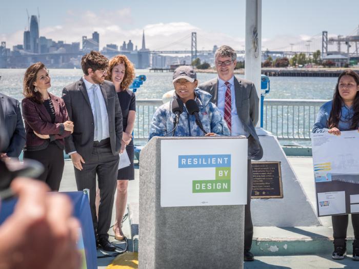 A speaker in a patterned jacket and baseball cap addresses a crowd from a white podium featuring the Resilient by Design logo. They are flanked by four colleagues on a waterfront pier, with one person holding a large design board. The San Francisco skyline and bay are visible in the background.