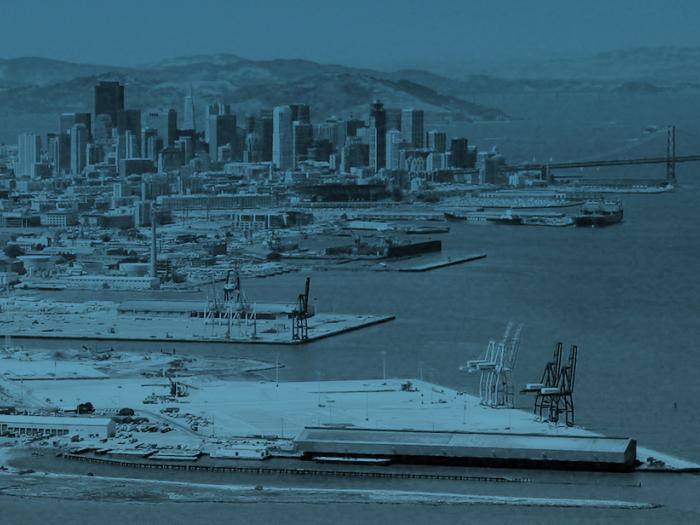 High-angle, blue-toned view of the San Francisco waterfront looking toward the downtown skyline and the Bay Bridge. Industrial piers with shipping cranes and a large warehouse occupy the foreground and middle ground, with dense urban skyscrapers and rolling hills in the distance.