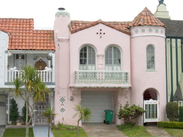 A row of three colorful, attached Mediterranean-style houses featuring pastel pink and beige facades, ornate arched windows, and decorative rooflines under a clear sky.