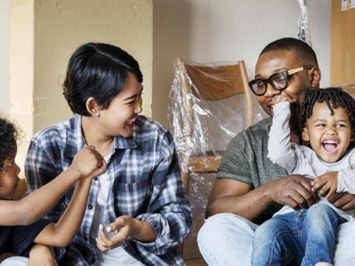 A happy family of three—a mother, father, and young child—celebrating in their new home. They are surrounded by cardboard moving boxes, with the father holding the child and everyone laughing together.