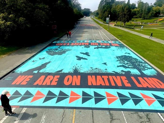 Mural on a road reading "We Are On Native Land" with nature silhouettes and geometric patterns.