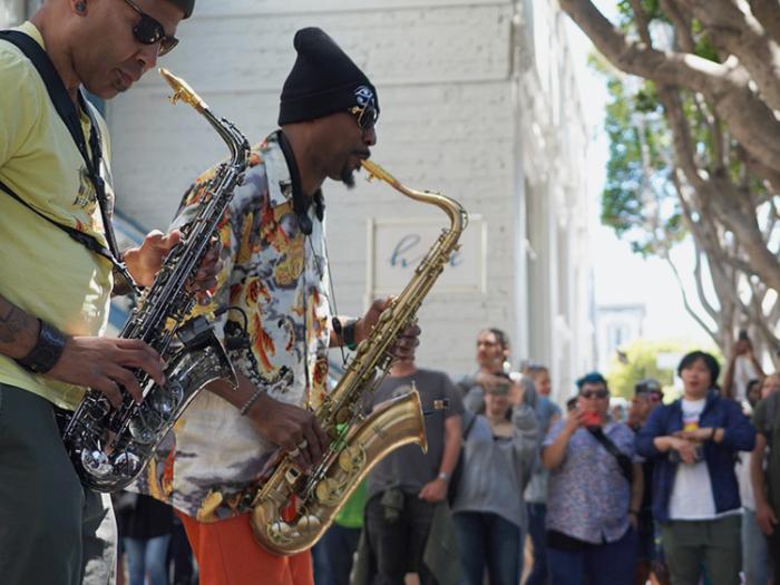 Two saxophone players in colorful clothes play a sidewalk concert for onlookers
