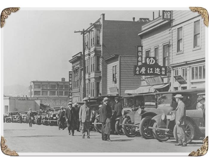 A black and white historical photograph of original Japantown in SoMa, from 1910 showing a street scene with several early 20th-century cars parked in front of multi-story wooden buildings. People in period clothing are standing on the sidewalk