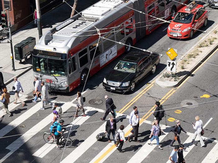 High-angle view of pedestrians in a crosswalk, with Muni bus, a black sedan and red taxi stopped at red light.