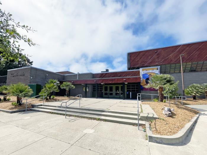 Exterior view of the Ella Hill Hutch Community Center, featuring a concrete and brown-paneled building facade, a wide concrete staircase leading up to the entrance, landscaping with small palm trees, and a partly cloudy blue sky.